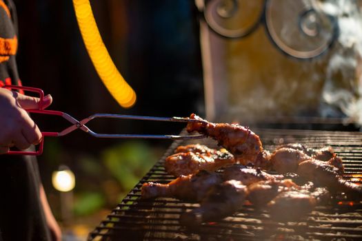human hand turns chicken drumsticks on a barbecue grill with grilling tongs. cooking food on an open fire in the evening. backyard party