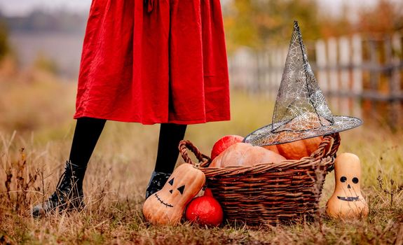 Halloween pumpkins and basket with witch hat standing at girls feet on autumn ground