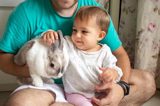 adorable baby sits in dad's arms and strokes a decorative rabbit. father shows little child easter bunny. domestic animals in a family with children