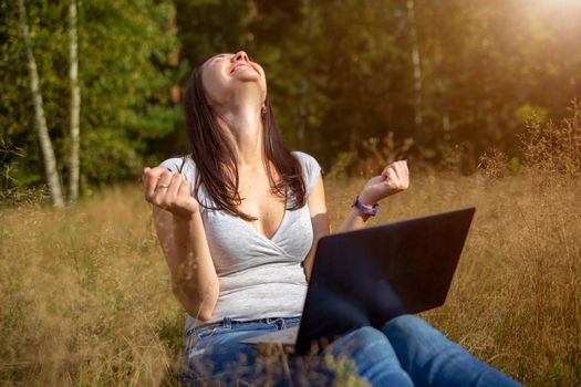 happy young woman with laptop on a sunny lawn. freelancer works in nature. student studies remotely on nature landscape outdoor