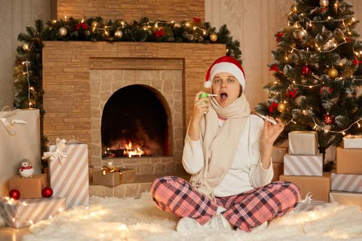 Woman having problem with health, being at home, lady sitting in floor with crossed legs, showing thermometer to camera, using sore throat spray, wearing home closing and santa hat.