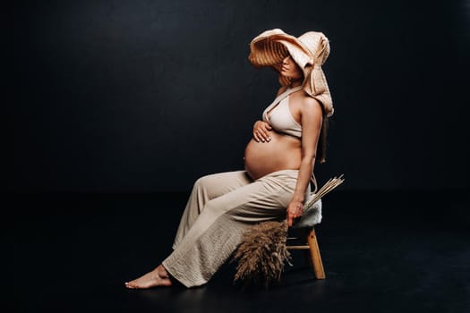 pregnant woman in a straw hat with beige clothes with a bouquet in her hands in the studio on a black background.