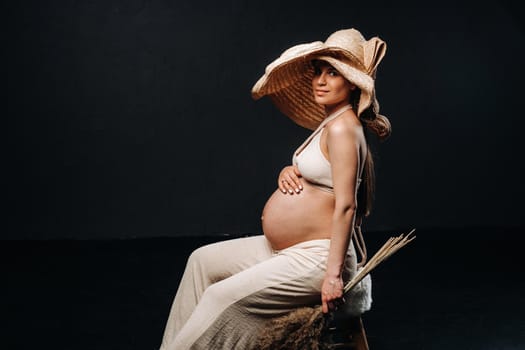 pregnant woman in a straw hat with beige clothes with a bouquet in her hands in the studio on a black background.