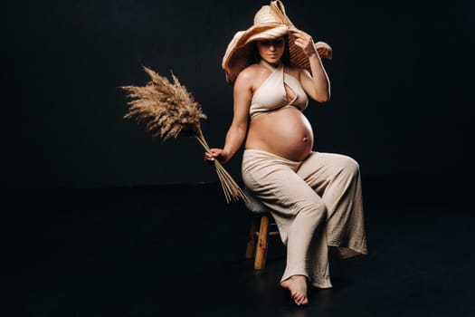 pregnant woman in a straw hat with beige clothes with a bouquet in her hands in the studio on a black background.