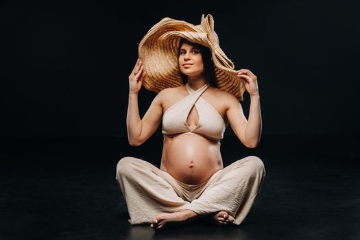 a pregnant woman in a straw hat sits on the floor in beige clothes in a studio on a black background.