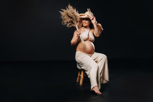 pregnant woman in a straw hat with beige clothes with a bouquet in her hands in the studio on a black background.