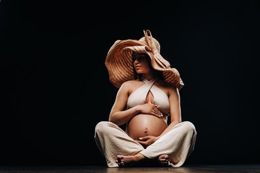 a pregnant woman in a straw hat sits on the floor in beige clothes in a studio on a black background.