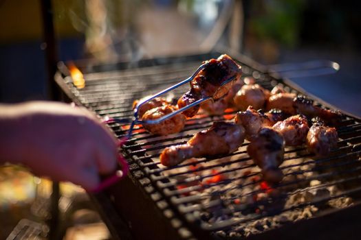 human hand turns chicken drumsticks on a barbecue grill with grilling tongs. cooking food on an open fire in the evening. backyard party