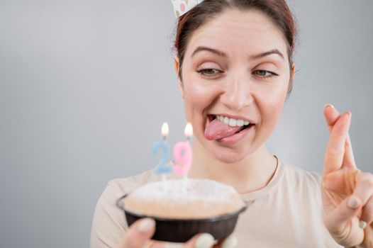 Happy caucasian woman sticking out her tongue and blowing out the candles on the cake with her fingers crossed. The girl celebrates her 29th birthday.