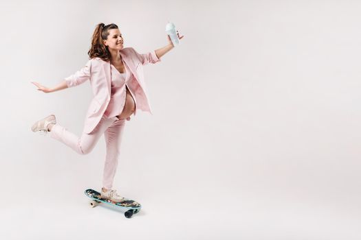 A pregnant girl in a pink suit with a skateboard in her hands stands on a gray background.