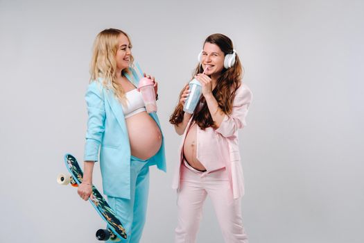 Two pregnant girls in turquoise and pink suits with glasses of juice, a skate and headphones stand on a gray background.