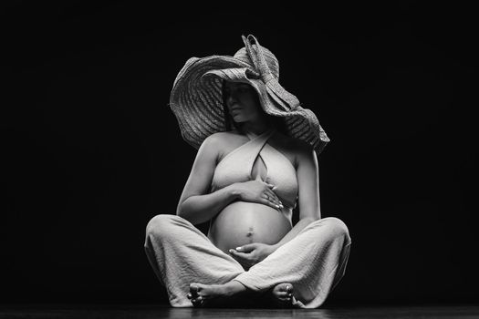 a pregnant woman in a straw hat sits on the floor in beige clothes in a studio on a black background.
