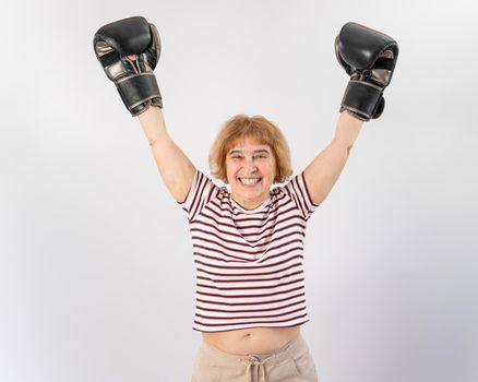 An elderly woman in fighting gloves raises her hands up on a white background. Victory in a duel.