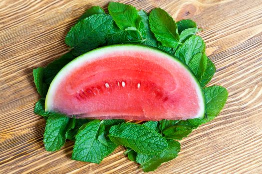 A red slice of watermelon lying on a wooden table. Around the berries lie the leaves of green mint. Photo close up