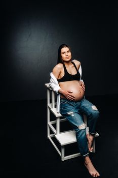 pregnant woman sitting on a ladder chair in a studio on a black background.