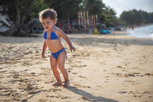 happy little girl in a swimsuit runs along the beach with palm trees. adorable toddler girl in bikini on sandy beach of tropical ocean in sunshine