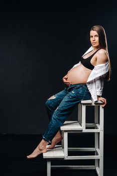 pregnant woman sitting on a ladder chair in a studio on a black background.