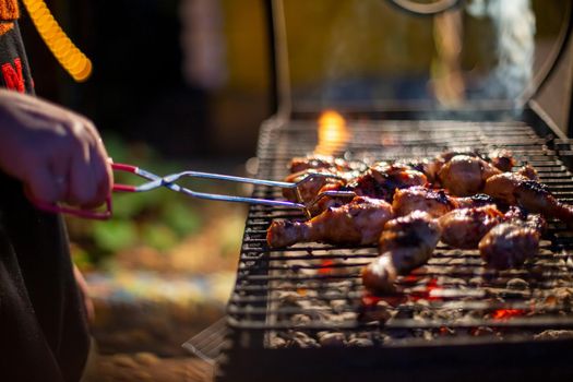 human hand turns chicken drumsticks on a barbecue grill with grilling tongs. cooking food on an open fire in the evening. backyard party