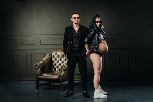 a pregnant woman in black clothes and a headscarf and a man in a suit in a studio on a dark background.