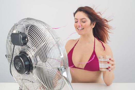 Red-haired smiling woman in a bikini drinks a cold drink and enjoys the blowing wind from an electric fan on a white background. Climate control on a hot summer day.