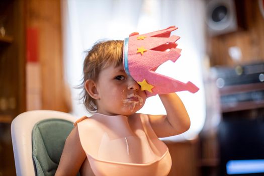 adorable toddler in a crown sits at home in a bib. little Princess with funny expression home interior