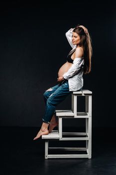 pregnant woman sitting on a ladder chair in a studio on a black background.