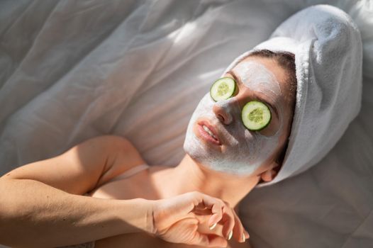 A woman with a towel on her hair and in a clay face mask and cucumbers in front of her eyes lies on the sheet.