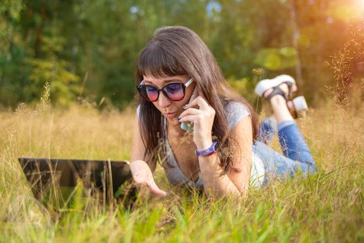 young woman swears on the phone while looking into a laptop. woman freelancer works in a sunny meadow