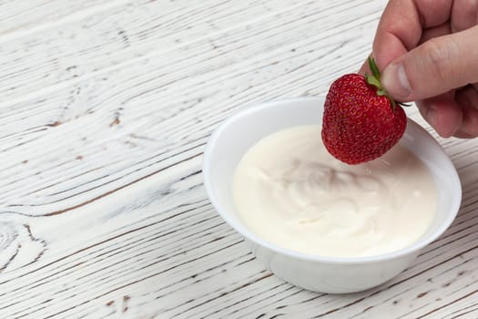 men's hand dips strawberries in sour cream on white wooden background