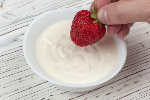 men's hand dips strawberries in sour cream on white wooden background