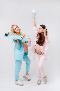 Two pregnant girls in turquoise and pink suits with glasses of juice, a skate and headphones stand on a gray background.