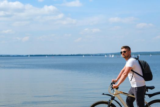 A cyclist with a backpack and glasses rides along the sea on a bicycle on a beautiful sunny day.