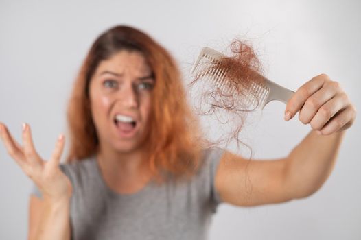 Caucasian woman with a grimace of horror holds a comb with a bun of hair. Hair loss and female alopecia