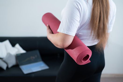 Young chubby woman folds sports mat after online fitness class.