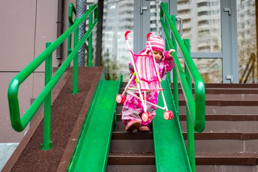 cute toddler with a toy stroller walks along steel railing ramp for wheelchair, carts and strollers. gentle descent from the stairs outdoors
