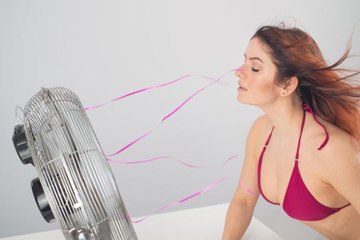 Red-haired smiling woman in a bikini drinks a cold drink and enjoys the blowing wind from an electric fan on a white background. Climate control on a hot summer day.