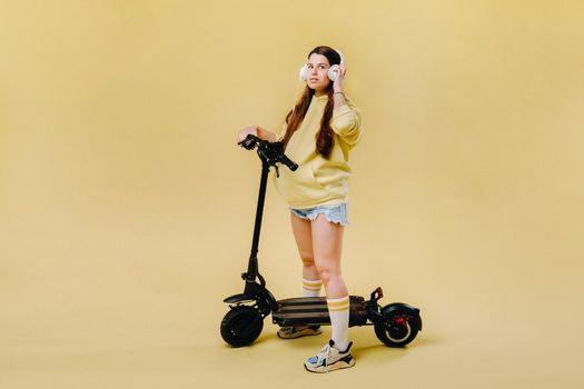 Pregnant girl in yellow clothes and headphones on an electric scooter on an isolated yellow background.