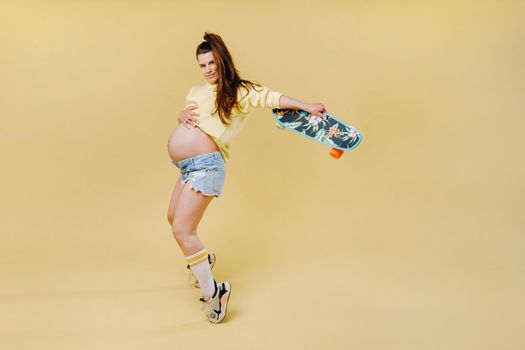 Pregnant girl in a yellow jacket with a skateboard in her hands on a yellow background.