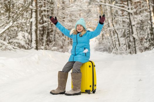 A girl in winter in felt boots sits on a suitcase on a frosty snowy day.