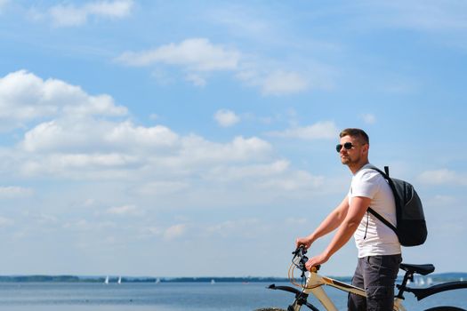 A cyclist with a backpack and glasses rides along the sea on a bicycle on a beautiful sunny day.