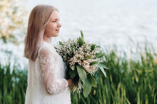 A beautiful nine-year-old blonde girl with long hair in a long white dress, holding a bouquet of lilies of the valley flowers, walking in nature in the Park.Summer, sunset