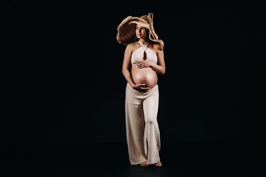 pregnant woman in a straw hat in beige clothes in the studio on a black background.