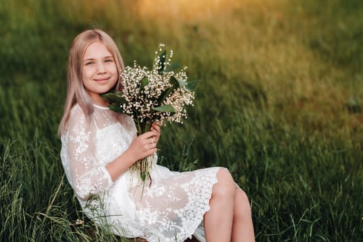 A beautiful nine-year-old blonde girl with long hair in a long white dress, holding a bouquet of lilies of the valley flowers, walking in nature in the Park.Summer, sunset