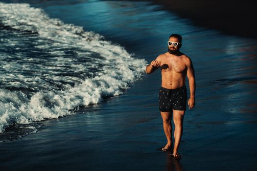 a strong man in black shorts and glasses walks along the beach with black volcanic sand on the island of Tenerife. Spain.