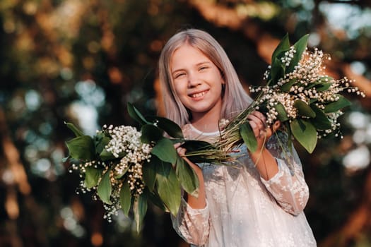 A beautiful nine-year-old blonde girl with long hair in a long white dress, holding a bouquet of lilies of the valley flowers, walking in nature in the Park.Summer, sunset