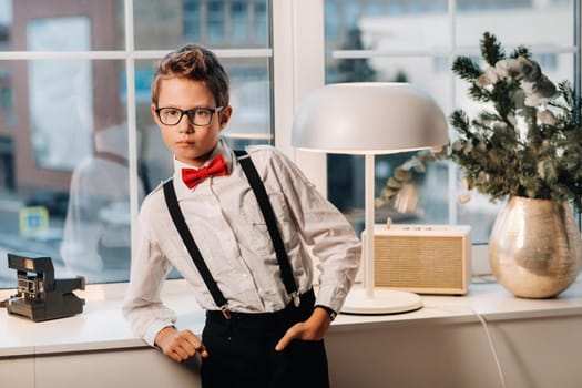 A boy in a shirt with a red bow tie and glasses stands at the window