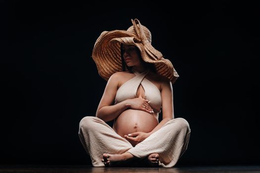 a pregnant woman in a straw hat sits on the floor in beige clothes in a studio on a black background.