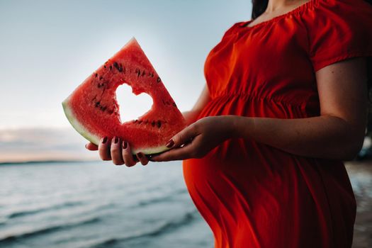 close-up of a pregnant woman holding a piece of watermelon with a heart made with it, standing in nature.