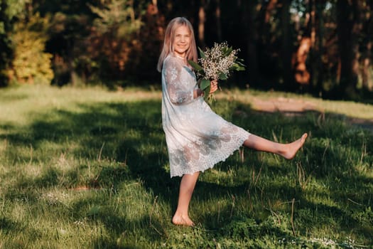 A beautiful nine-year-old blonde girl with long hair in a long white dress, holding a bouquet of lilies of the valley flowers, walking in nature in the Park.Summer, sunset