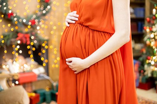 best christmas gift. faceless pregnant woman in a red dress hugs her belly against the background of the Christmas living room in blur. close-up no face.